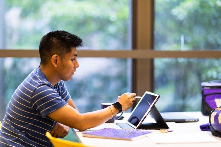 A student studies at a table with a tablet and notebook in a bright campus space overlooking greenery.